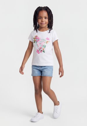 Jeune fille aux cheveux tressés portant un t-shirt blanc à motif floral, un short en jean bleu clair et des baskets blanches, debout sur un fond blanc.
