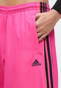 Rózsaszín sportnadrág elasztikus derékpánttal, oldalzsebbel és fekete háromcsíkkal díszítve, valamint fekete Adidas logóval.