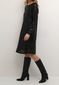 Kvinna som går, iklädd en knälång svart paljettklänning med långa ärmar och svarta klackade knähöga stövlar mot en enkel bakgrund.