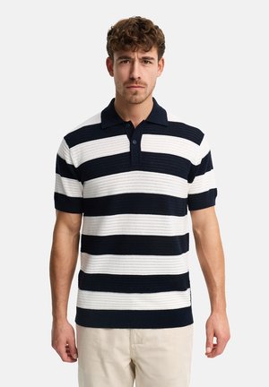 Uomo con capelli ricci che indossa una polo a maniche corte a righe blu navy e bianco e pantaloni beige chiaro, in piedi davanti a uno sfondo semplice.