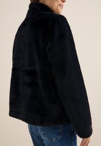Veste en peluche noire avec un col haut, présentant une texture douce et une coupe décontractée, associée à un jean en denim bleu en arrière-plan.
