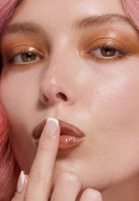 Nærbillede af et ansigt med pink hår, bronzefarvet øjenskygge og glansfulde brune læber. Finger placeret nær læberne, korte nude negle med hvide spidser.