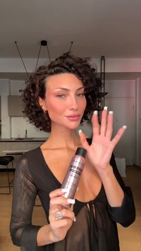 Hårprodukt i en rosa flaska med svart kork, märkt "Superfix." Modell med lockigt hår, iförd en svart genomskinlig topp, håller upp flaskan.