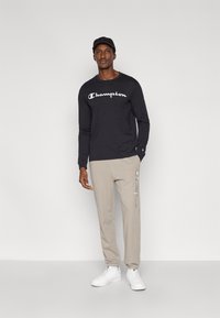 Μαύρο μακρυμάνικο T-shirt με λευκό λογότυπο "Champion", συνδυασμένο με ανοιχτό μπεζ φόρμα με πλαϊνό λογότυπο και λευκά αθλητικά παπούτσια.