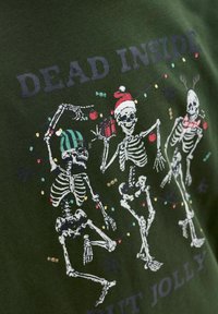 Trois squelettes dansants portant des accessoires de Noël et des lumières avec le texte "Mort à l'intérieur mais joyeux" sur un tissu vert foncé.