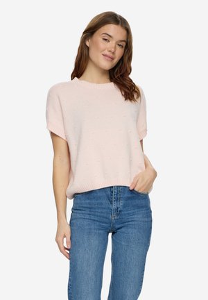 Giovane donna che indossa un maglione a maniche corte di colore rosa chiaro e jeans blu a vita alta, in piedi con una mano in tasca su sfondo bianco.