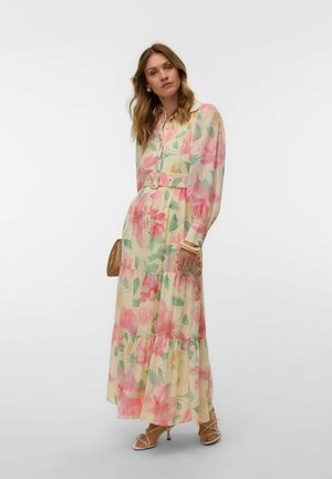 Kvinde iført en lang, bæltepræget blomsterkjole med pink, grønne og gule mønstre, kombineret med snørede høje hæle og holder en lille brun håndtaske.