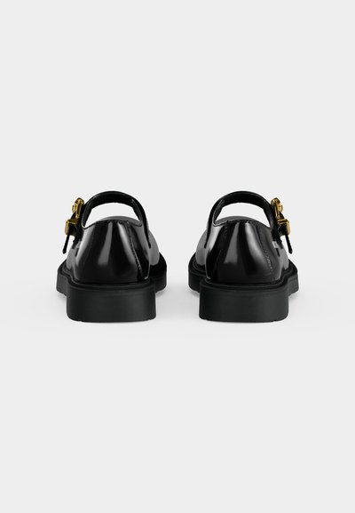 Paire de chaussures Mary Jane en cuir noir avec semelles basses et épaisses et boucles dorées, vues de dos sur un fond clair uni.