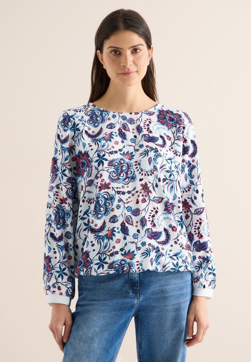 Femme portant un chemisier blanc à manches longues avec des motifs floraux bleus et violets, associée à un jean bleu, debout devant un fond uni.