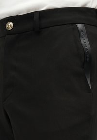 Μαύρο παντελόνι με χρυσό κουμπί και διακοσμητικό πλαϊνή τσέπη με ανάγλυφο λογότυπο "Versace Jeans Couture".