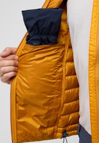 Giacca isolante arancione con una texture liscia e lucida. Presenta una tasca interna che contiene un oggetto di tessuto blu piegato. Chiusura frontale con zip, dettaglio elastico.