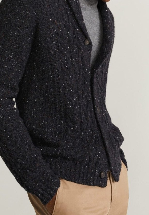 Homme portant un cardigan à torsades bleu marine foncé avec des boutons, par-dessus une chemise grise et un pantalon beige, les mains dans les poches, sur un fond uni.