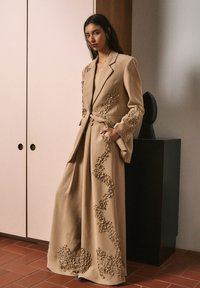 Blazer beige sur mesure et pantalon à jambes larges avec broderie florale le long des bords. Tissu lisse avec une silhouette structurée.