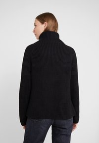Black ribbed sweater with a high collar, long sleeves, and a subtle split at the back hem. Worn with dark jeans.