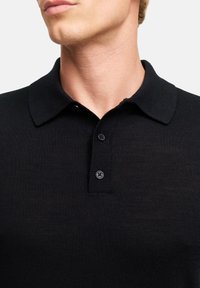 Polo shirt nera a coste con colletto classico, dotata di una patta con tre bottoni e tessuto testurizzato, presentata su uno sfondo neutro.