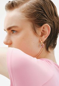 Pendientes de aro de oro con acentos en mármol rosa. Primer plano de una oreja, con cabello corto y una camiseta rosa claro. Textura de piel suave.