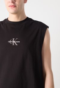Juoda be rankovių paminkštinta marškinėliai iš medvilnės, su apvaliu iškirpimu ir sidabro spalvos siuvinėtu Calvin Klein Jeans logotipu priekyje.