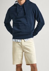 Marinblå hoodie med kenguruficka och broderad logotyp, tillsammans med beige shorts. Ljusrandig skjorta synlig vid kragen.