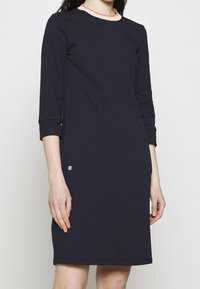 Marindress med långa ärmar, rund halsringning som har en ljusrosa och gräddfärgad kant, samt en liten logodetalj på den nedre sidan. Slät textur.