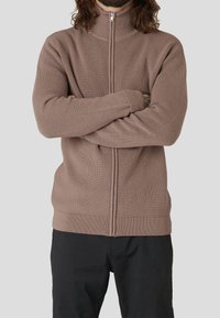 Beiger Zip-Up-Pullover mit gerippter Textur und hohem Kragen, ausgestattet mit langen Ärmeln und einer taillierten Passform, kombiniert mit schwarzen Hosen.