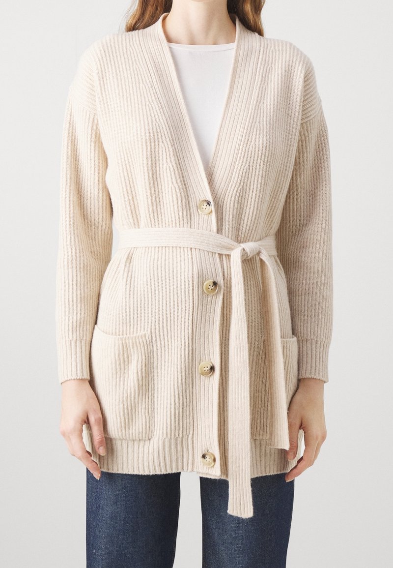 Femme portant un cardigan en maille côtelée crème avec ceinture nouée, fermeture à boutons, poches devant et manches longues, sur une chemise blanche et un jean bleu.