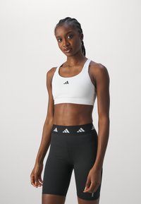 Vit idrottsbehå med rund halsringning och svart logotyp. Svarta höga shorts med vit märkesresår. Slät tygtextur.