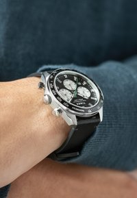Schwarze Chronographuhr mit silbernen Akzenten, Gehäuse aus Edelstahl, schwarzem Gummiband, weißen Subdials und einer tachymetrischen Skalenlünette.