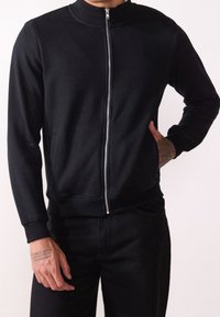 Felpa nera con zip, colletto alto, polsini e orlo a costine. Presenta una texture liscia e tasche laterali, indossata sopra pantaloni neri.