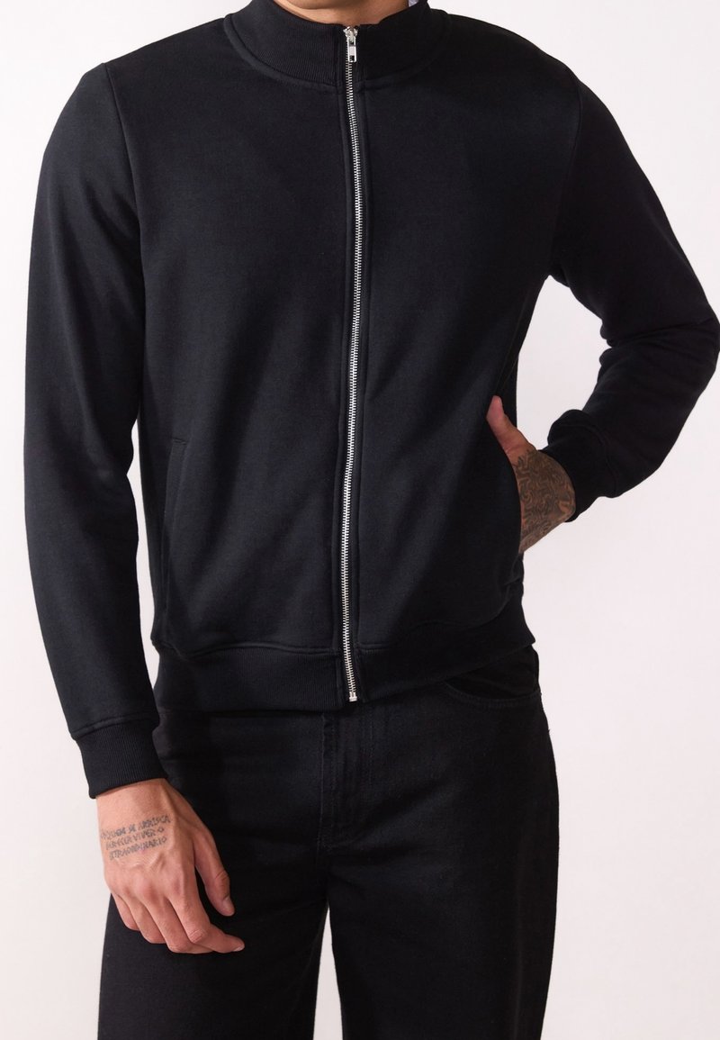 Felpa nera con zip, colletto alto, polsini e orlo a costine. Presenta una texture liscia e tasche laterali, indossata sopra pantaloni neri.