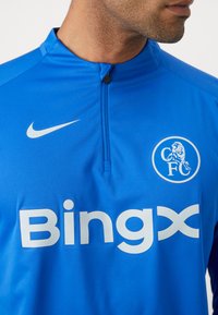 Blå Nike sportstrøje med kvart lynlås, der har et stort hvidt "Bingx" logo og et cirkulært hvidt "CFC" emblem på brystet. Glat tekstur.