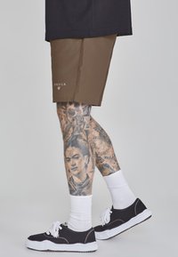 Shorts marron avec détails de logo, baskets blanches avec accents noirs, et chaussettes blanches. Tatouages détaillés visibles sur les jambes.
