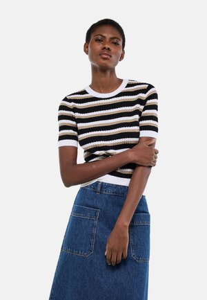 Femme aux cheveux courts portant un pull à manches courtes rayé noir, blanc et beige, ainsi qu'une jupe en denim bleu avec des poches devant.