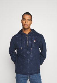 Felpa con zip di colore blu navy, dotata di cappuccio con cordoncino, caratterizzata da un delicato motivo a diamante e un dettaglio del logo sul lato sinistro del petto. Tessuto morbido.