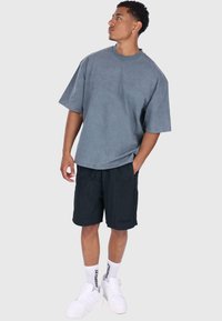 Graues, übergroßes T-Shirt mit Rundhalsausschnitt, kombiniert mit schwarzen Shorts. Weiße klobige Sneaker und Socken mit schwarzem Schriftzug runden das Outfit ab.