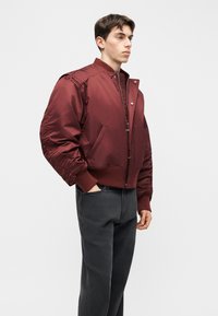 Νεαρός άνδρας που φοράει μπορντό bomber μπουφάν με ριμπ μανσέτες και μαύρα τζιν, στέκεται με το ένα χέρι στην τσέπη μπροστά σε απλό φόντο.