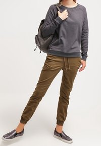 Felpa grigia con polsini a costine, pantaloni jogger marroni a sigaretta con caviglie elastiche e scarpe slip-on metalliche con dettagli testurizzati.