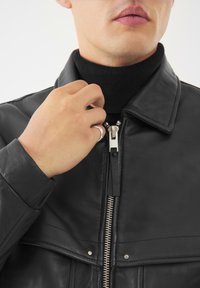 Giacca di pelle nera con zip davanti, colletto piegato e dettagli di cucitura visibili. Mano che tiene il colletto, indossando un dolcevita nero sotto.