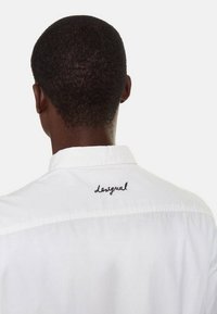 Osoba s krátkými tmavými vlasy, oblékající bílou košili s černým vyšitým nápisem "Desigual" na horní části zad pod límcem.