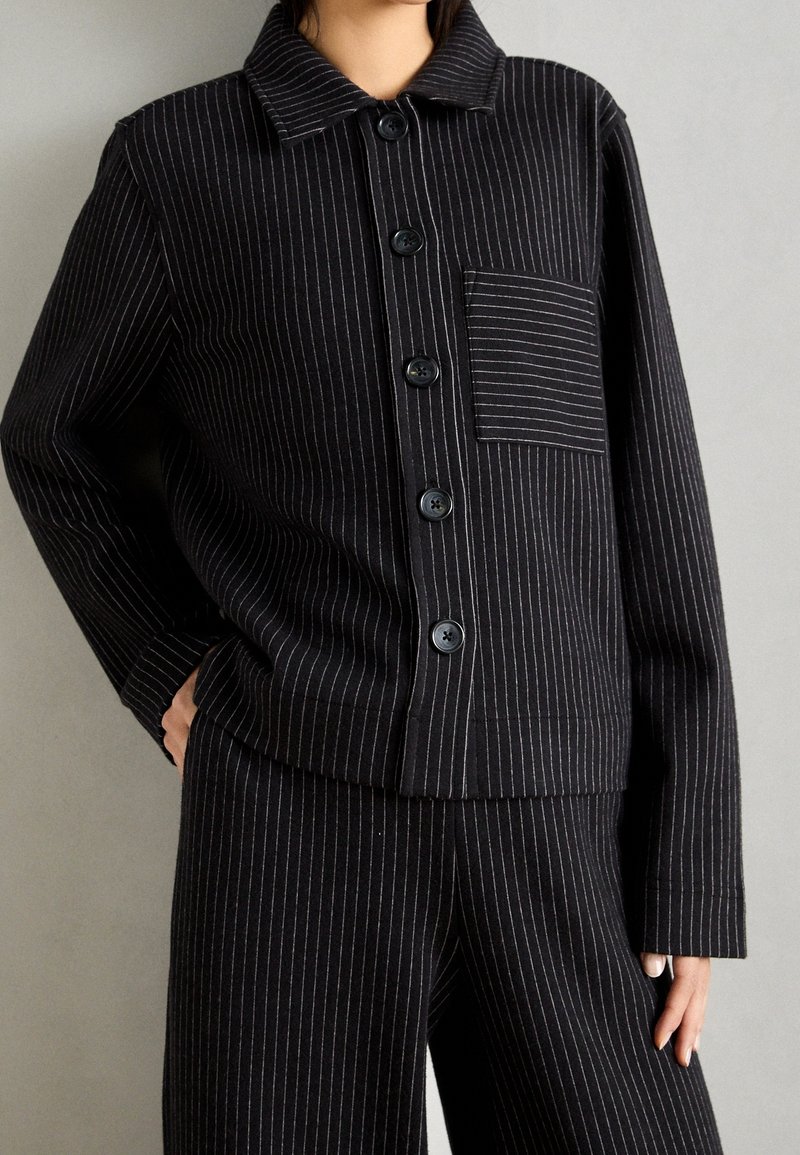 Czarna marynarka w prążki z kołnierzykiem, zapinana na guziki z przodu, z kieszenią naszywaną. Tkanina wydaje się strukturalna. Pasujące spodnie w prążki.