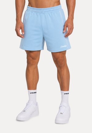 Man draagt lichtblauwe sportshorts, witte crew-sokken met tekst en witte sneakers, staand tegen een effen achtergrond.