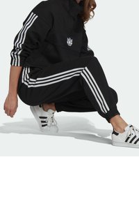 Person som bär en svart Adidas träningsdräkt med vita ränder och vita Adidas sneakers, hukande på en ljus bakgrund.
