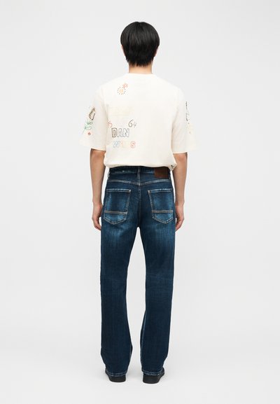 Άτομο που στέκεται με την πλάτη, φορώντας κεντημένο λευκό πουκάμισο με κείμενο και λουλούδια, συνδυασμένο με μπλε τζιν και μαύρα παπούτσια.