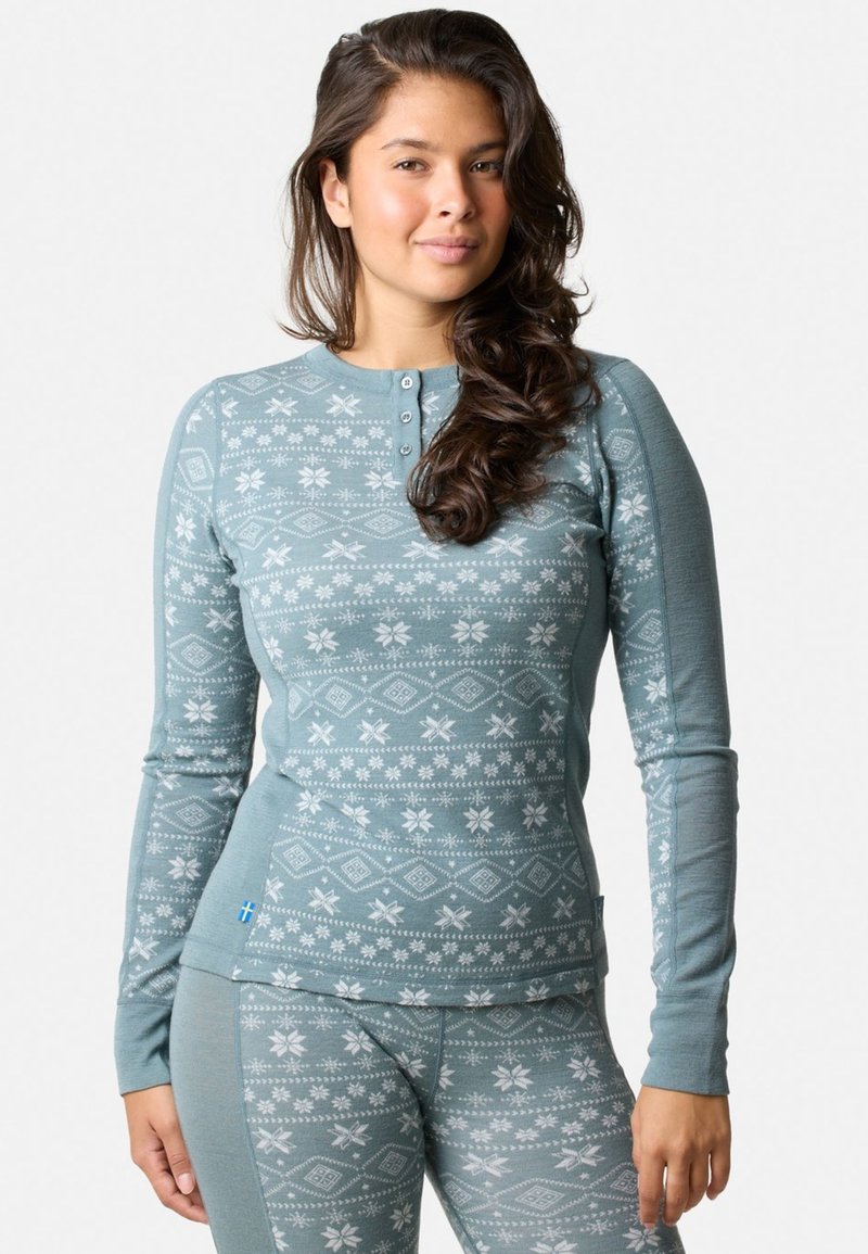 Langdurige mouwtop in teal met een wit sneeuwvlokpatroon, met knopen bij de halslijn en een getailleerd ontwerp. Zacht materiaal en een gestructureerde afwerking.