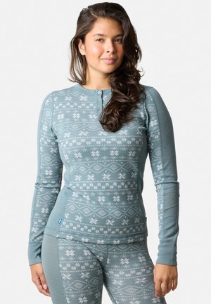 En ung kvinde med langt bølget hår iført en matchende blå mønstret langærmet top og leggings med snefnugsdesign.