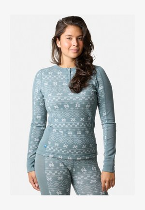 Giovane donna con lunghi capelli ondulati, che indossa un top a maniche lunghe e leggings coordinati di colore blu con motivi di fiocchi di neve.