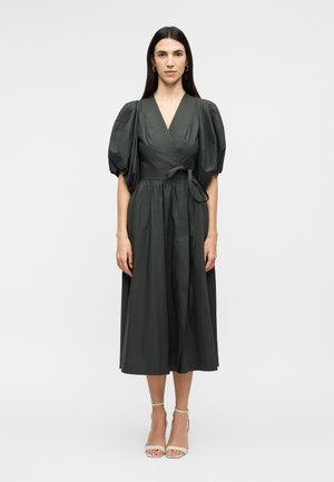 Moteris su ilgais tamsiais plaukais, vilkinti tamsiai žalią midi ilgio suknelę su pūstomis trumpomis rankovėmis ir dėvinti smėlio spalvos aukštakulnius, stovinti prie vientisos spalvos fono.