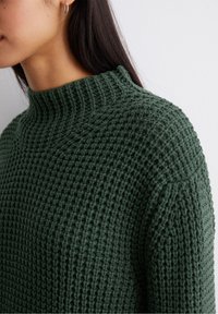 Ciemnozielony dziany sweter z wysokim prążkowanym kołnierzem i teksturowanym wzorem, o luźnym kroju i widocznym prążkowaniu na ramionach.