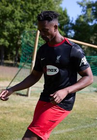 Jersey deportivo negro con acentos rojos y el logo del Liverpool FC; combinado con pantalones cortos rojos. El material parece ligero y que absorbe la humedad.