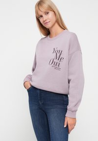 Jasnofioletowy sweatshirt z okrągłym dekoltem, z kontrastowym napisem w ciemnym brązie. Miękka tekstura, luźny krój i prążkowane mankiety.