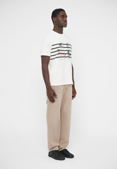 Λευκό κοντομάνικο μπλουζάκι με γραφικό σχέδιο σε μαύρες και κόκκινες ρίγες, σε συνδυασμό με μπεζ cargo παντελόνι φαρδιάς γραμμής και μαύρα αθλητικά παπούτσια, πλάγια όψη.